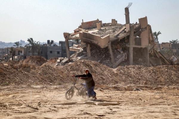 A man pushes a bicycle along a heavily damaged road past a destroyed building in Khan Yunis